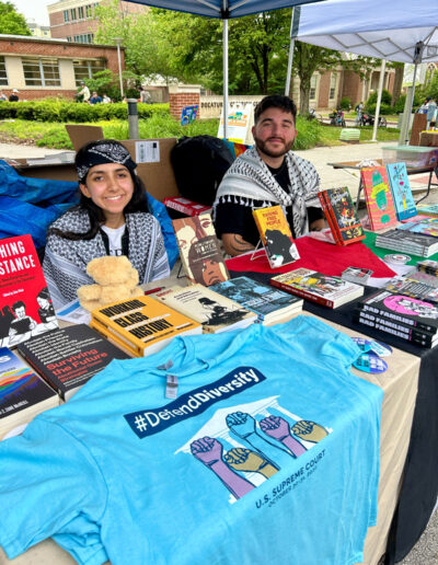 A couple of people at a booth with multiple books and a t-shirt on display.