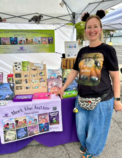 Gina Gallois, an author wearing a shirt that says “I'm the author” at the festival, smiling at her booth in front of her books.